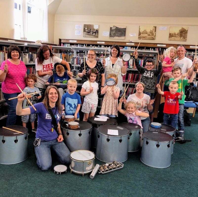 Community drum workshop in a library, group smiles for finale picture
